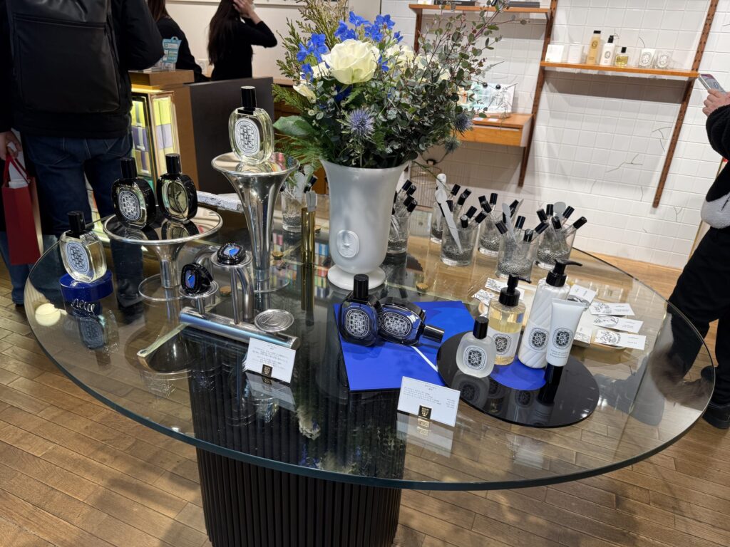 ガラスの円形テーブルに展示されたディプティック「オルフェオン」の香水コレクションと青と白のフラワーアレンジメント