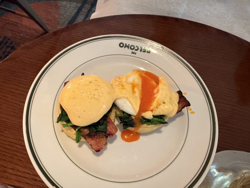 Two Eggs Benedict served on a white plate with THE BELCOMO logo. The right poached egg is cut open, revealing a rich orange yolk flowing over bacon, spinach, and a muffin.