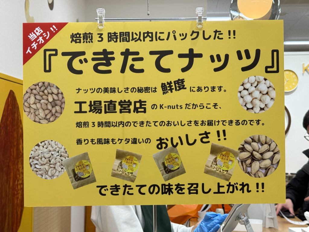 A bright yellow store sign advertising 'Freshly Made Nuts' packed within 3 hours of roasting at the factory-direct shop K-nuts, featuring photos of almonds, macadamias, cashews, and pistachios.