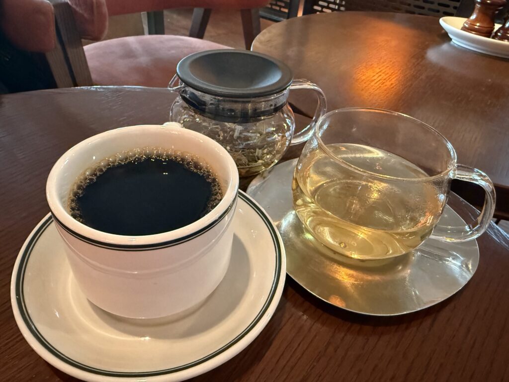 木目のテーブル上の白いカップに入ったブラックコーヒーと、ガラスのティーセットに入ったハーブティー