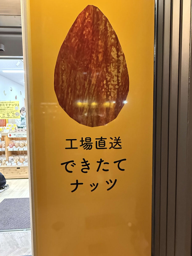 鮮やかな黄色の背景に大きなアーモンドの画像と、「工場直送 できたてナッツ」という黒い文字が記された店頭の看板。左側にはガラス越しに商品が並ぶ店内の様子が見える。