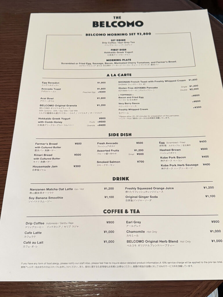 The breakfast menu for THE BELCOMO restaurant placed on a wooden table, listing details for the Morning Set, A La Carte items, Side Dishes, and Drinks.