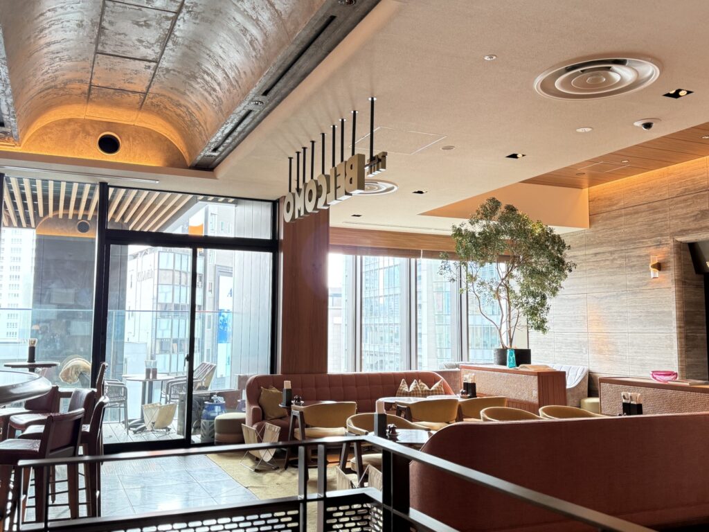 青山グランドホテル「THE BELCOMO」の店内。大きな窓から自然光が入り、テラコッタ色のソファや特徴的な天井の吊り看板、観葉植物が配置された開放的なラウンジ空間。