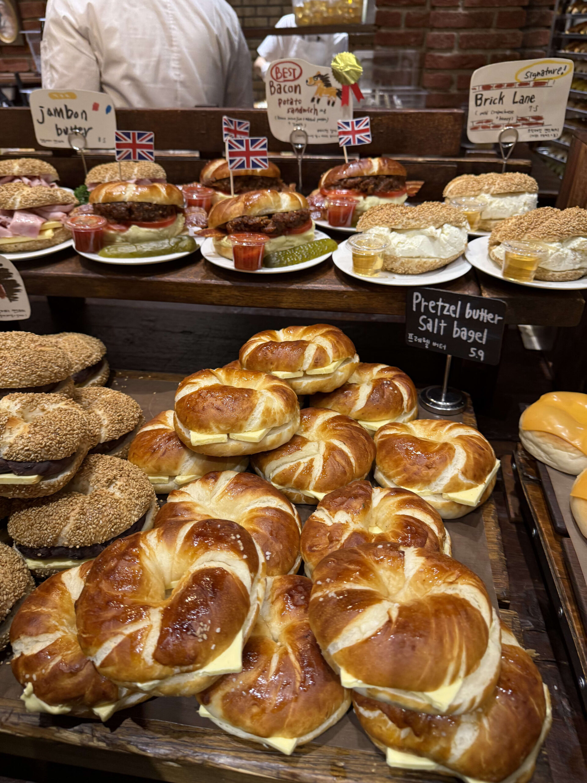 Pretzel bagels with butter and stuffed sandwiches displayed at a bakery.