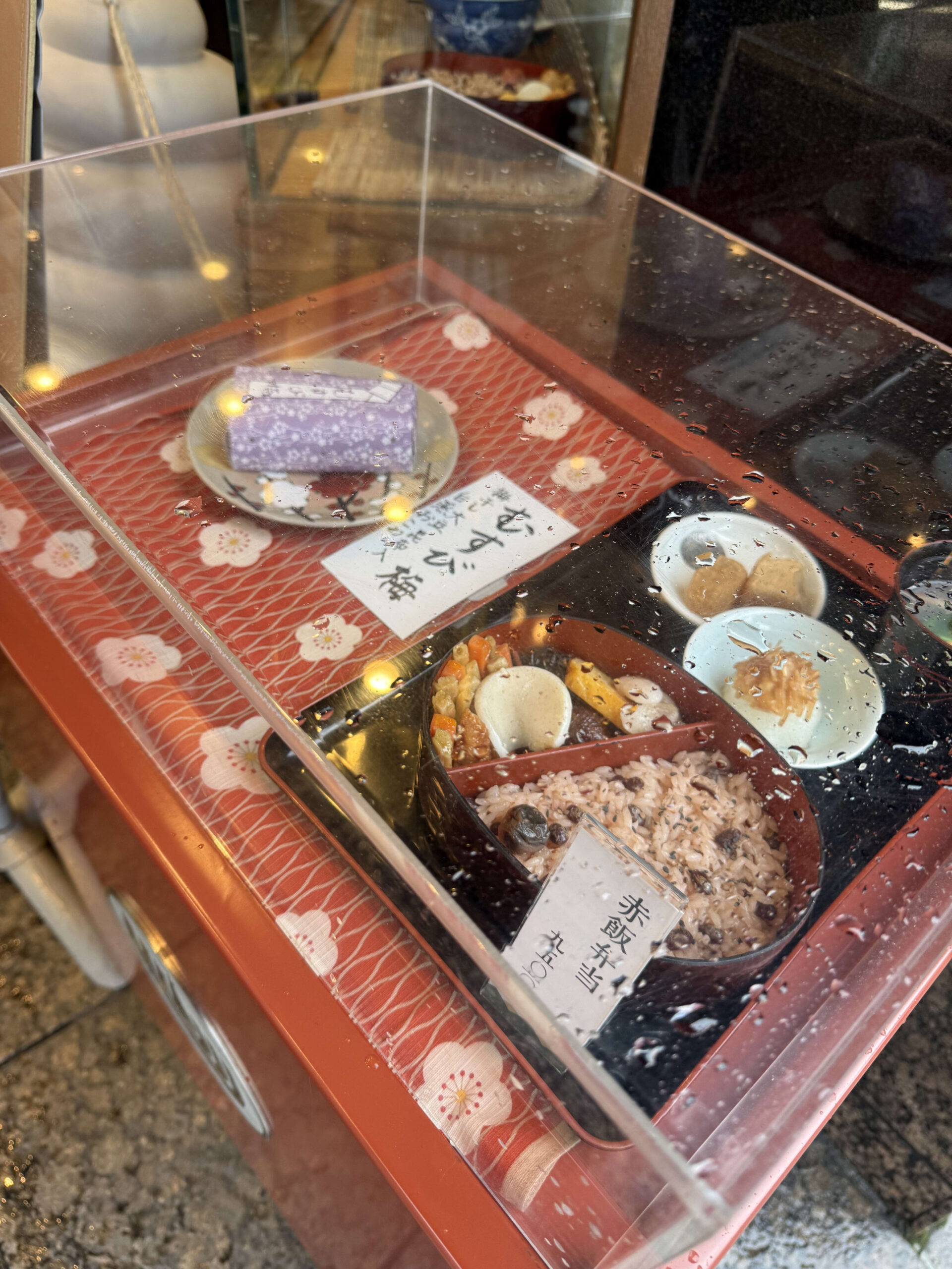雨粒がつくケースに並ぶ、老舗和菓子屋のむすび梅と赤飯弁当のサンプル。