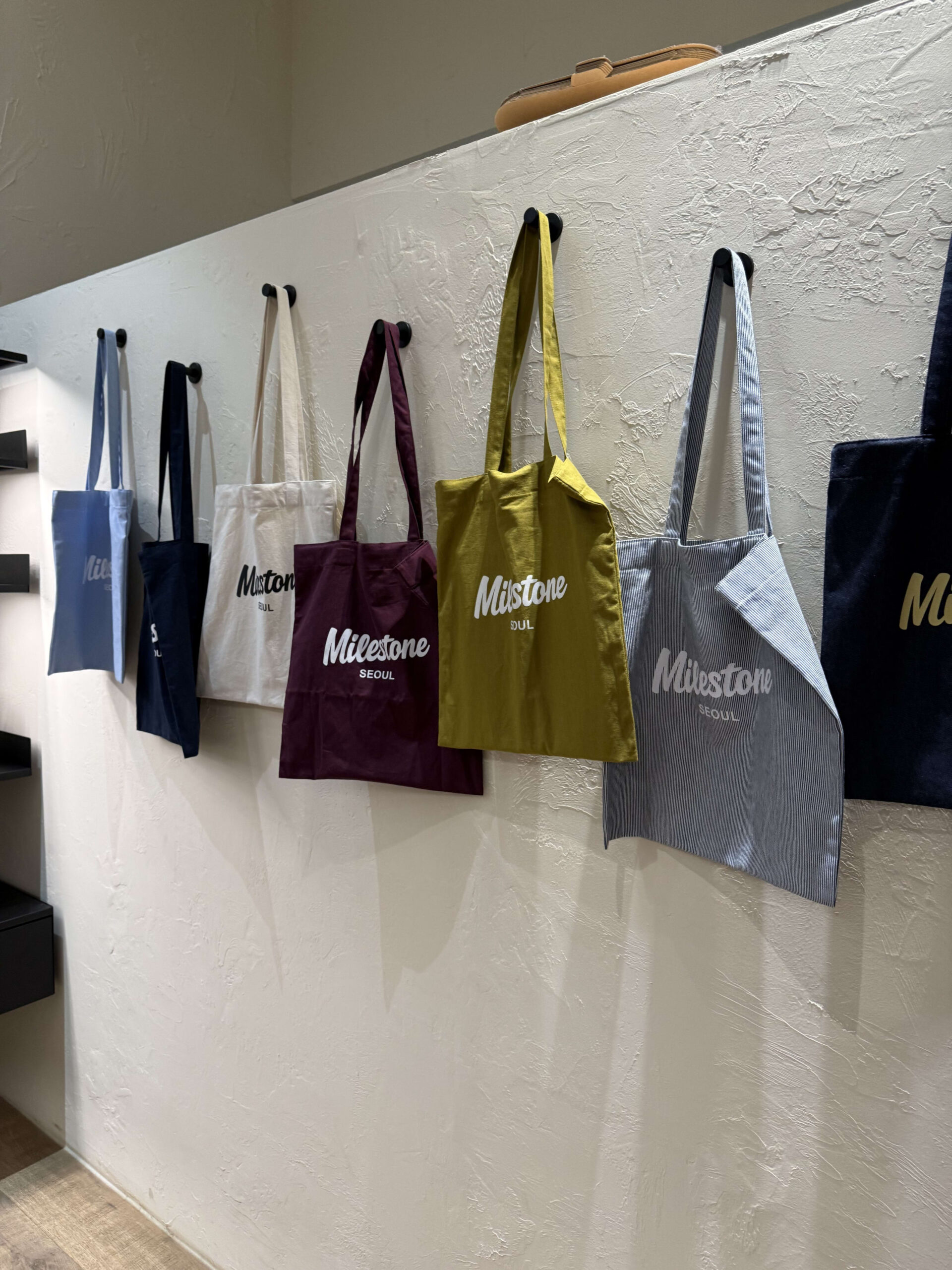 Colorful 'Milestone SEOUL' tote bags on a white wall.