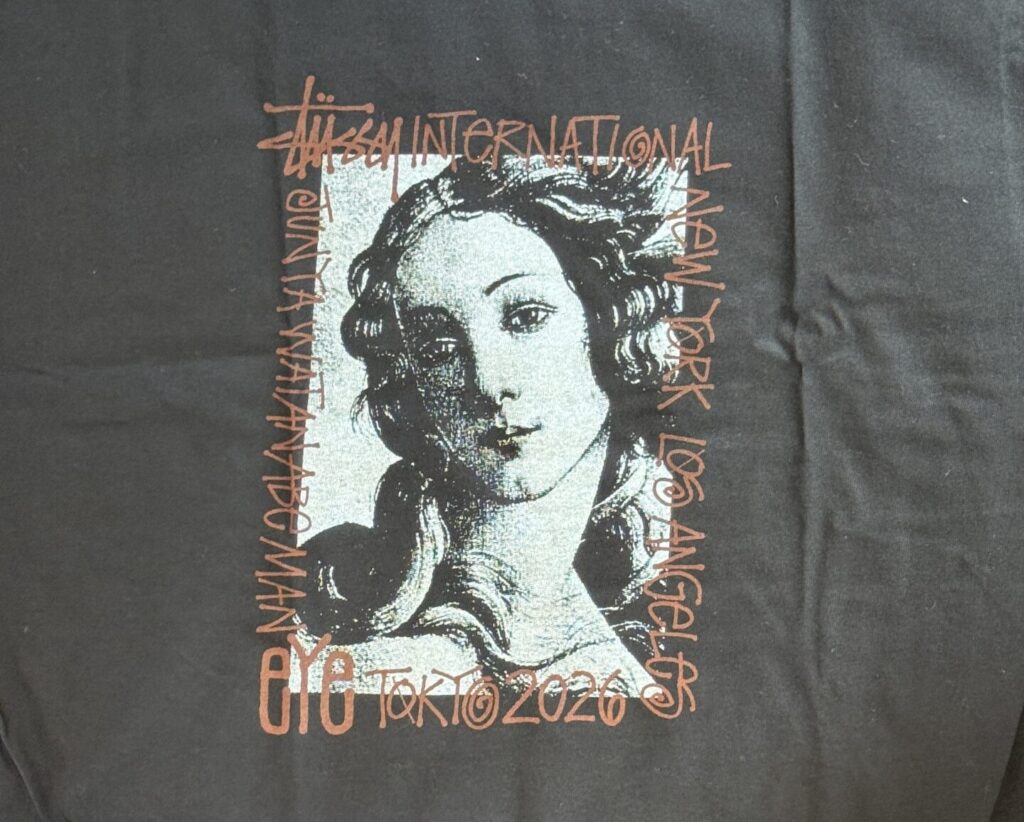 黒いTシャツの背面に配置された、eYe JUNYA WATANABE MAN x Stüssyコラボレーションのグラフィック。中央の女性の肖像画を囲むように、「STÜSSY INTERNATIONAL」や「eYe JUNYA WATANABE MAN」、「NEW YORK LOS ANGELES TOKYO 2026」のテキストが茶色い文字でプリントされている。