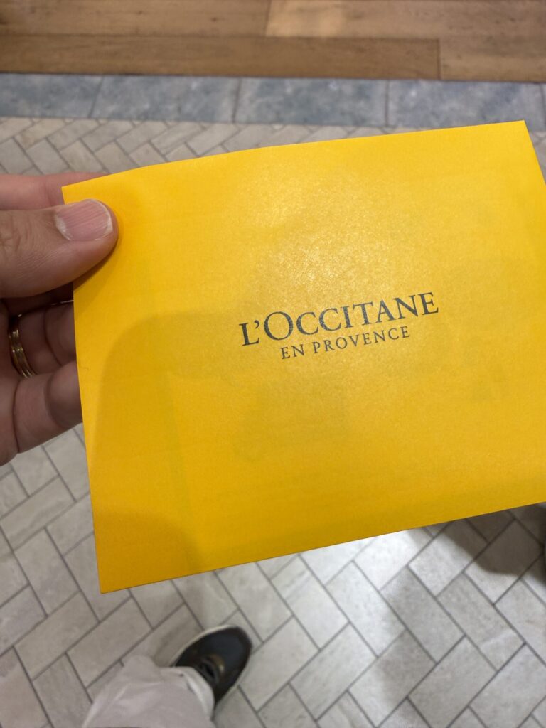A hand holding a bright yellow L'OCCITANE reservation envelope against a tiled floor background.