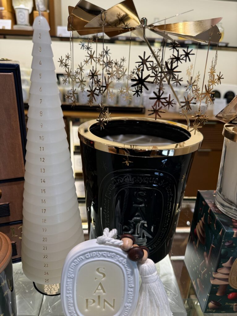 艶やかな漆黒のガラスにゴールドの縁取りが施された巨大なキャンドル。そこに刻まれた「SAPIN」の文字を見た瞬間、冬の澄んだ空気とモミの木の青々しい香りが鼻腔をくすぐるような錯覚に陥る。キャンドルの熱でくるくると回るであろうゴールドのカルーセルには繊細な星々が瞬き、ホリデー特有の華やかな温度感を放っている。手前に横たわるのは、1から25までの数字が静かに刻まれた純白のツリー型キャンドル。アドベントカレンダーの箱を開けるワクワク感とはまた違う、毎日少しずつ火を灯し、香りと共にクリスマスへのカウントダウンを刻むという、なんとも贅沢でロマンチックな時間の使い方の提案だ。傍らに添えられた白いオーバル型のオーナメントも、タッセルとウッドビーズのディテールが今年のテーマである「魔法の森」の温もりをさりげなく体現している。奥に覗く深いグリーンのパッケージが、昨年のゴールド基調から一転した今年のシックで愛らしい世界観を裏付けている。店舗に足を踏み入れ、このディスプレイを前にした時の「今年もこの季節が来た」という高揚感。いち早くこの空間に招かれた喜びと、これから始まるホリデーシーズンへの期待が、この一枚にぎゅっと凝縮されている。