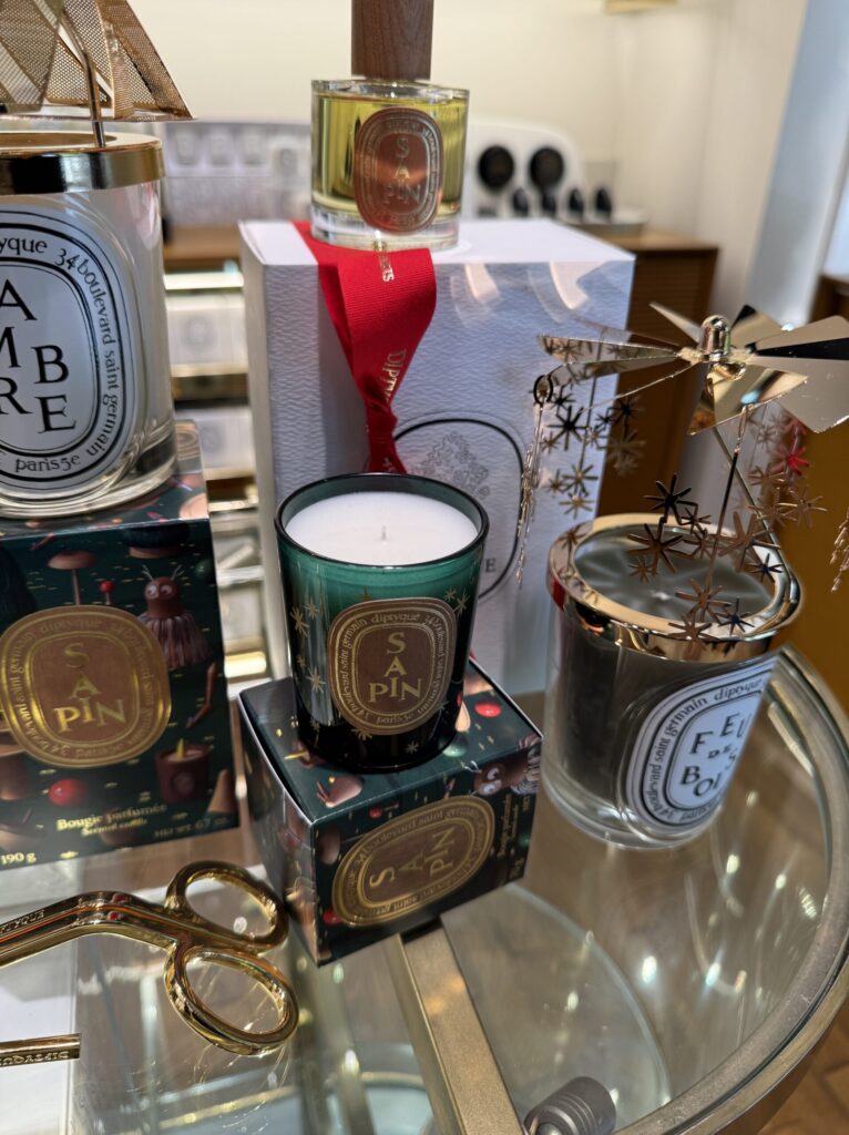 ガラスのテーブルに並べられたdiptyqueのホリデーコレクション。今年のテーマ「魔法の森」を象徴する深いグリーンの「Sapin（サパン）」キャンドルが、ひときわ目を引く。パッケージに描かれた愛らしいイラストからは、ジャン＝シモン・ロッシュが手掛けた木の温もりと遊び心が伝わってくる。火を灯す前から、冬の澄んだ空気とモミの木の清々しい香りがふわりと漂ってきそうだ。隣には、炎の熱でくるくると回るゴールドのカルーセルを纏ったキャンドルたち。繊細な星のモチーフが光を反射し、ホリデーならではの高揚感を煽る。赤いリボンが結ばれた純白のギフトボックスや、重厚なゴールドのウィックトリマーといったディテール一つひとつが、特別な季節の到来を告げている。昨年は完売で涙を呑んだアドベントカレンダーへの期待が、この美しいディスプレイを前にして確信へと変わる。サパンの香りに包まれながら、毎日少しずつ新しい香りと出会う至福のカウントダウン。この森の魔法に、今年もどっぷりと沼る準備はできている。