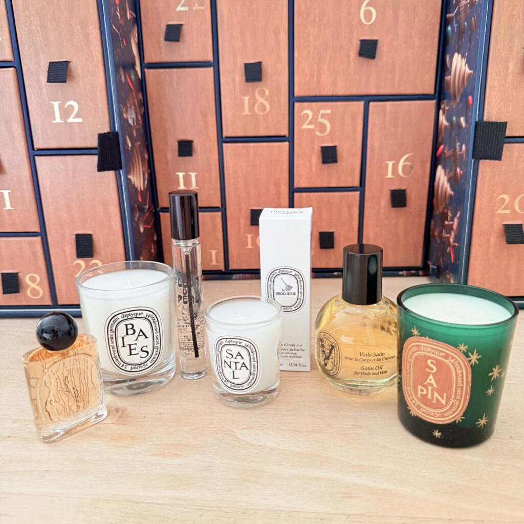 木の温もりを感じる引き出しが整然と並ぶ、diptyqueの2026年アドベントカレンダー。昨年買い逃した悔しさを晴らすように、今年は無事に手に入れた安堵と高揚感がこの一枚に詰まっている。背景にそびえるのは、ジャン＝シモン・ロッシュが手掛けた『魔法の森』の世界。その手前には、これから始まるホリデーのカウントダウンを彩る主役たちが誇らしげに並ぶ。右端でひと際目を引く深いグリーンのグラスは、限定キャンドル「Sapin（サパン）」。火を灯す前から、冬の澄んだ空気とモミの木の清々しい香りが漂ってくるようだ。左端には、高級感溢れる「Bois Corsé（ボワコルセ）」のミニボトル。香りの余韻が長く続くこのパルファンは、冬のお出かけの特別な相棒になるだろう。中央には定番人気の「Baies」や「Santal」のキャンドル、そして肌に寄り添う「Fleur de Peau」のオードパルファンが控える。さらに、今や貴重となった黄金色に輝く「サテンオイル」や、日常の空気を瞬時にリセットしてくれるバジルのルームスプレーの姿も。毎日ひとつずつ引き出しを開け、新しい香りと出会う。そんな贅沢な時間を少しずつ味わっていく期待感に胸が高鳴る。視覚からも嗅覚からも、ホリデーシーズンの魔法にどっぷりと沼る準備は万端だ。